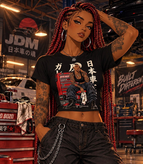 A woman with long red-and-black braids stands in a garage-like workshop wearing a black cropped graphic T-shirt with a red bus and Japanese text, paired with dark jeans and a silver chain accessory.